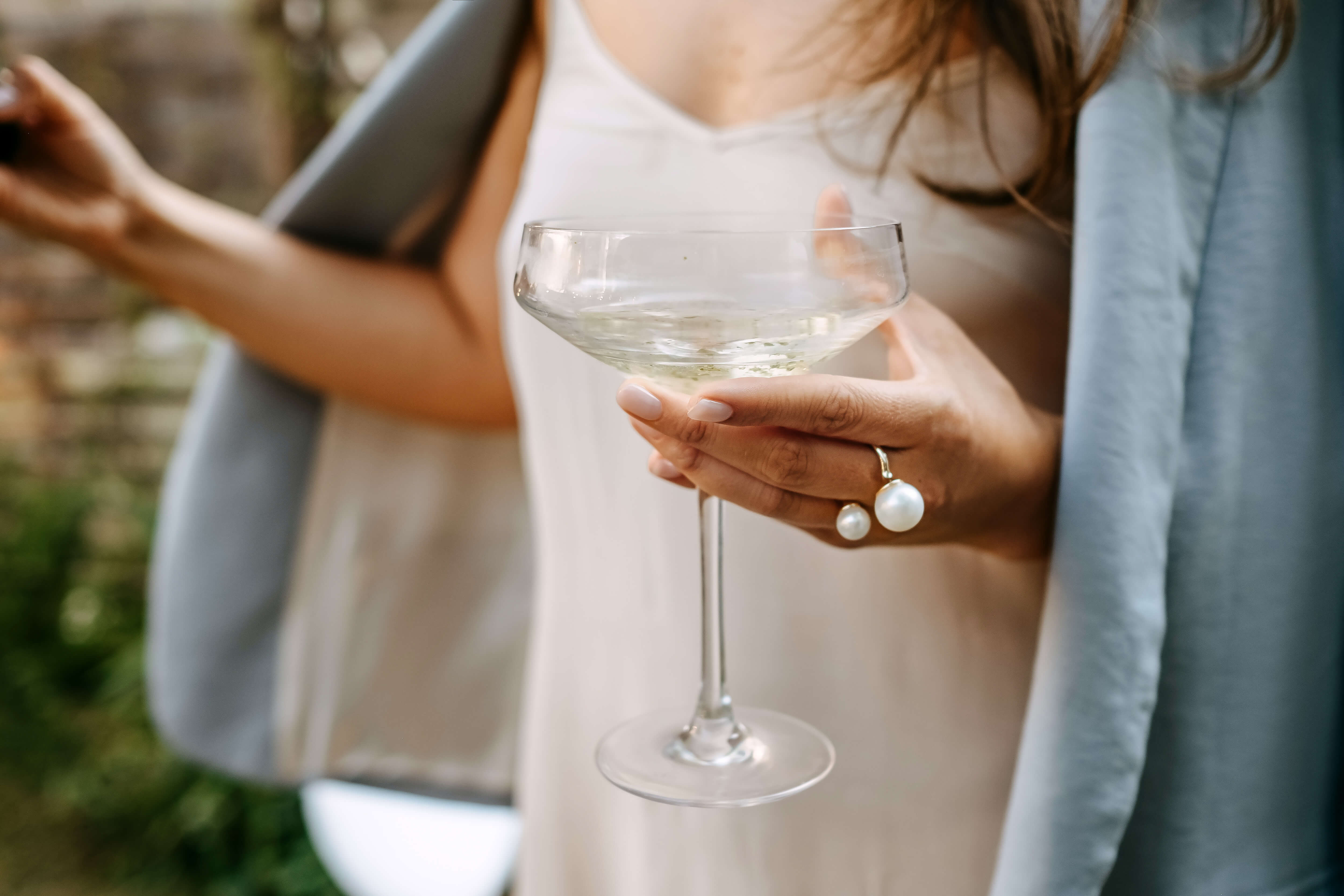 Frau hält ein Glas Pure Poetry Vodka bei einem Abendessen im Freien Eine Frau in eleganter Kleidung hält ein Glas Pure Poetry Vodka, während sie an einem festlich gedeckten Tisch im Freien sitzt. Auf dem Tisch sind Teller, Besteck, Zitronen und andere Dekorationen zu sehen.