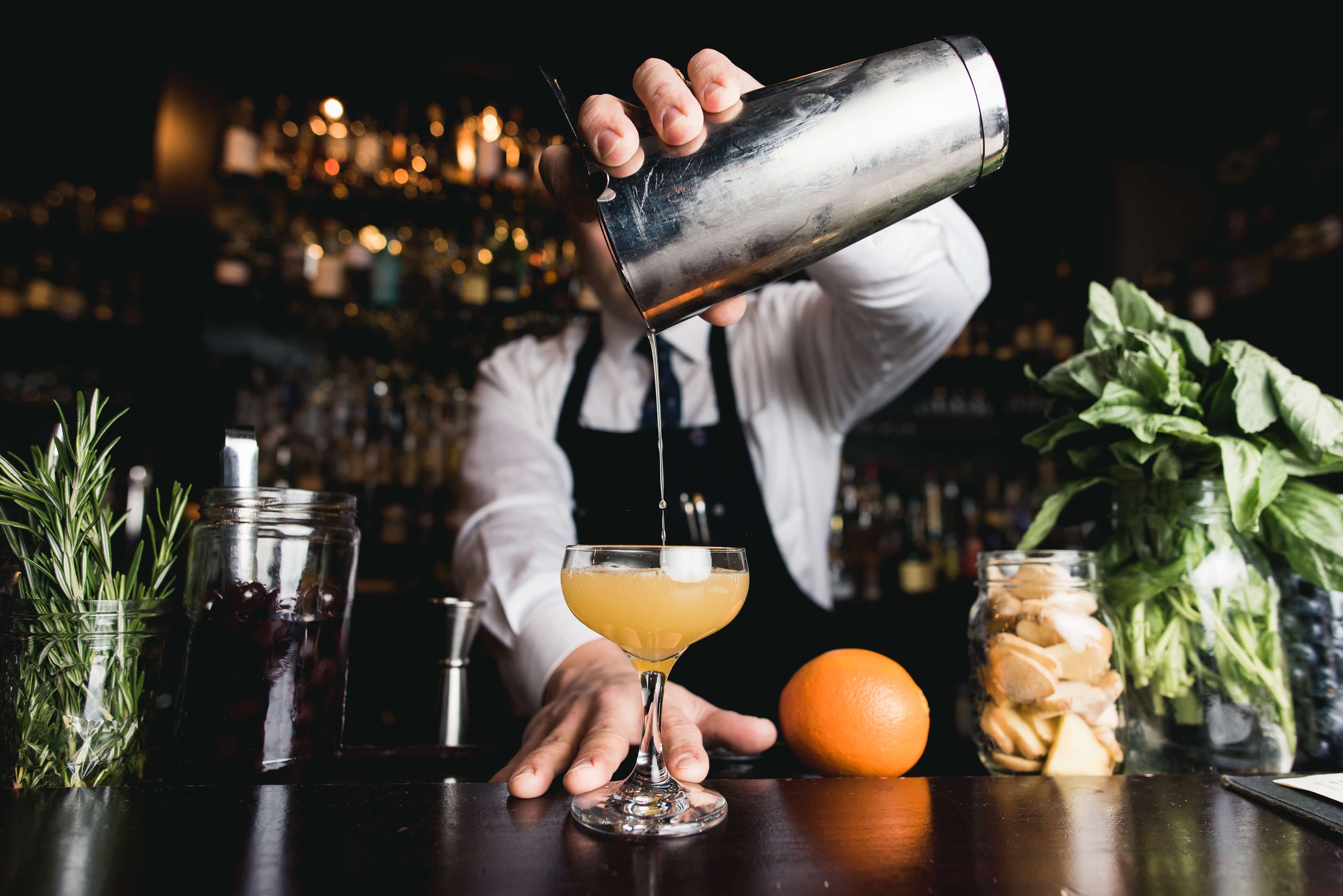 Ein Barkeeper in einer Bar gießt mit einem Cocktailshaker eine gelbe Flüssigkeit in ein Cocktailglas. Im Vordergrund sind frische Kräuter, eine Orange und Einmachgläser mit Zutaten zu sehen. Barkeeper mixt einen Cocktail mit Poetry Pure Vokda
