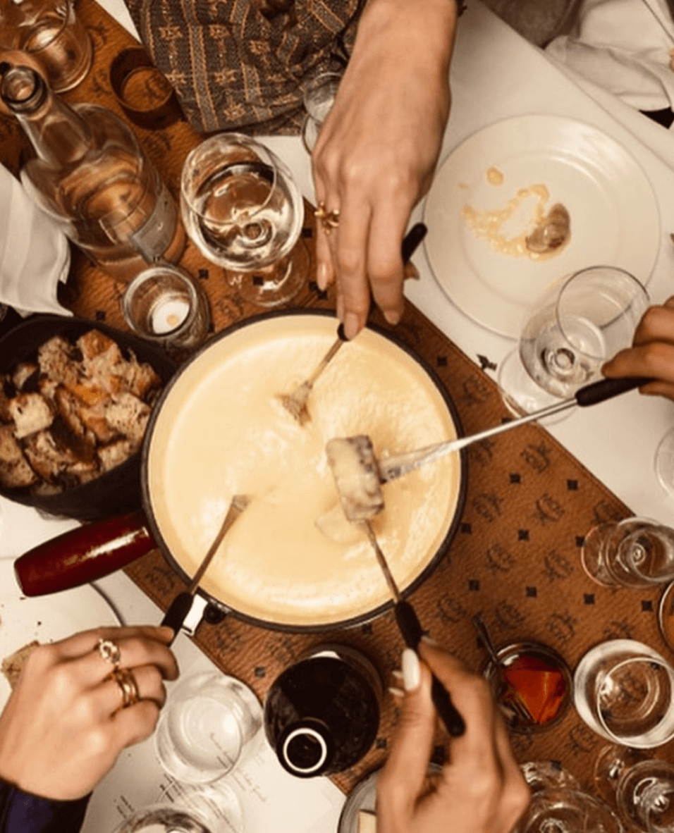 Eine Gruppe von Freunden versammelt sich um einen Tisch, um ein leckeres Käsefondue zu genießen. Die Szene zeigt Hände, die Brot in den geschmolzenen Käse tauchen, umgeben von verschiedenen Getränken. Die gemütliche Atmosphäre ist ideal für gesellige Aben