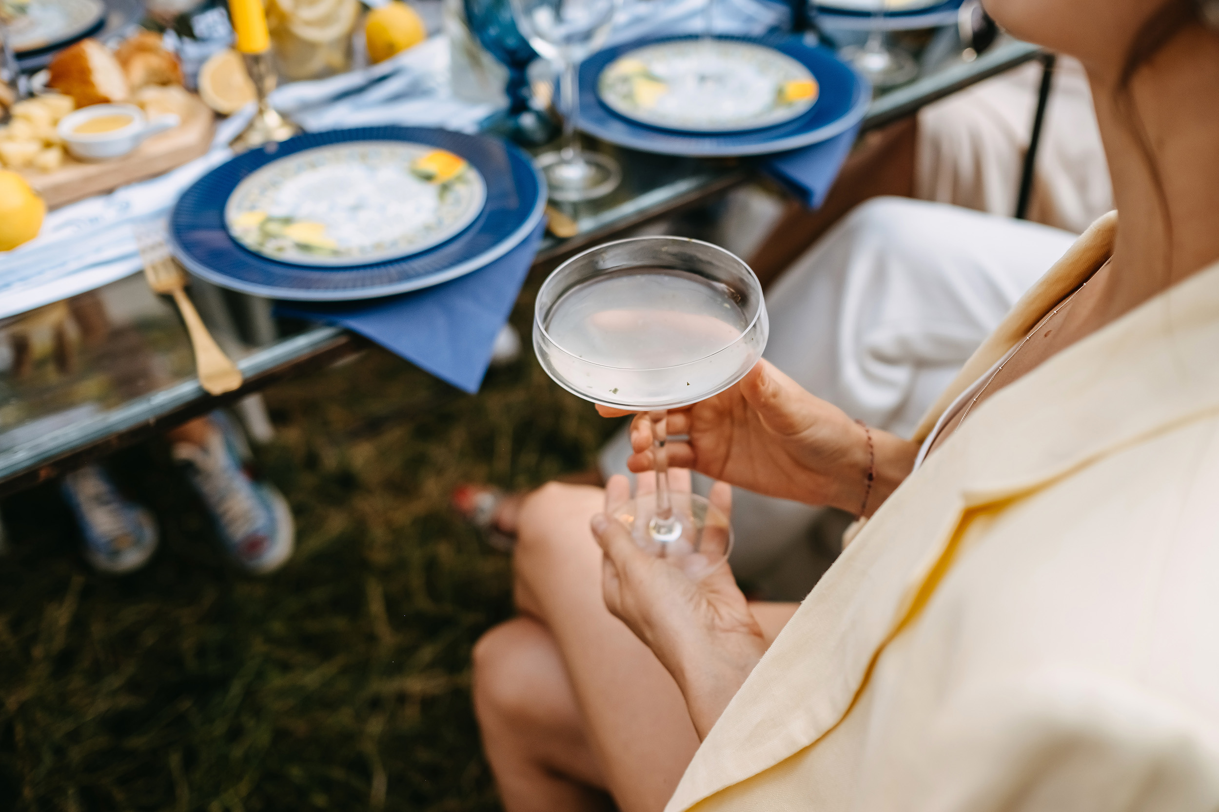 Eine Frau in einem weißen Kleid und einer hellen Jacke hält ein elegantes Cocktailglas mit Pure Poetry Vodka. Im Hintergrund sind Pflanzen und unscharfe Elemente zu sehen, die auf eine Gartenparty hindeuten.